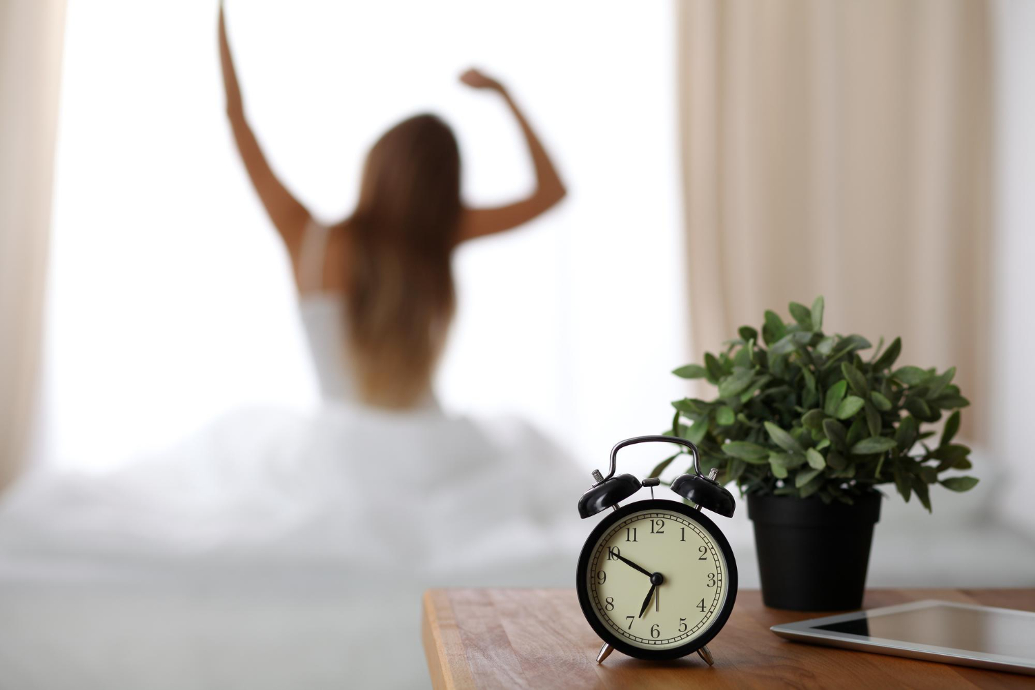 A clock on a side table and a woman waking up in the background